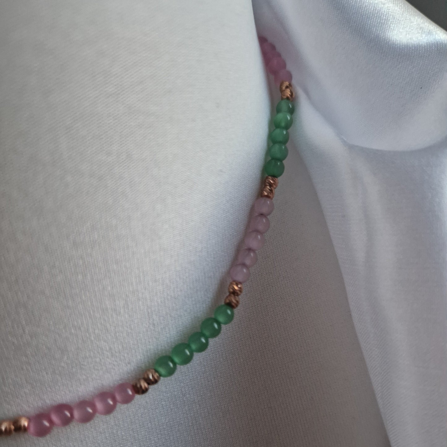 Collana di perline rosa e verdi con dettagli in metallo color rame, su sfondo di tessuto bianco.