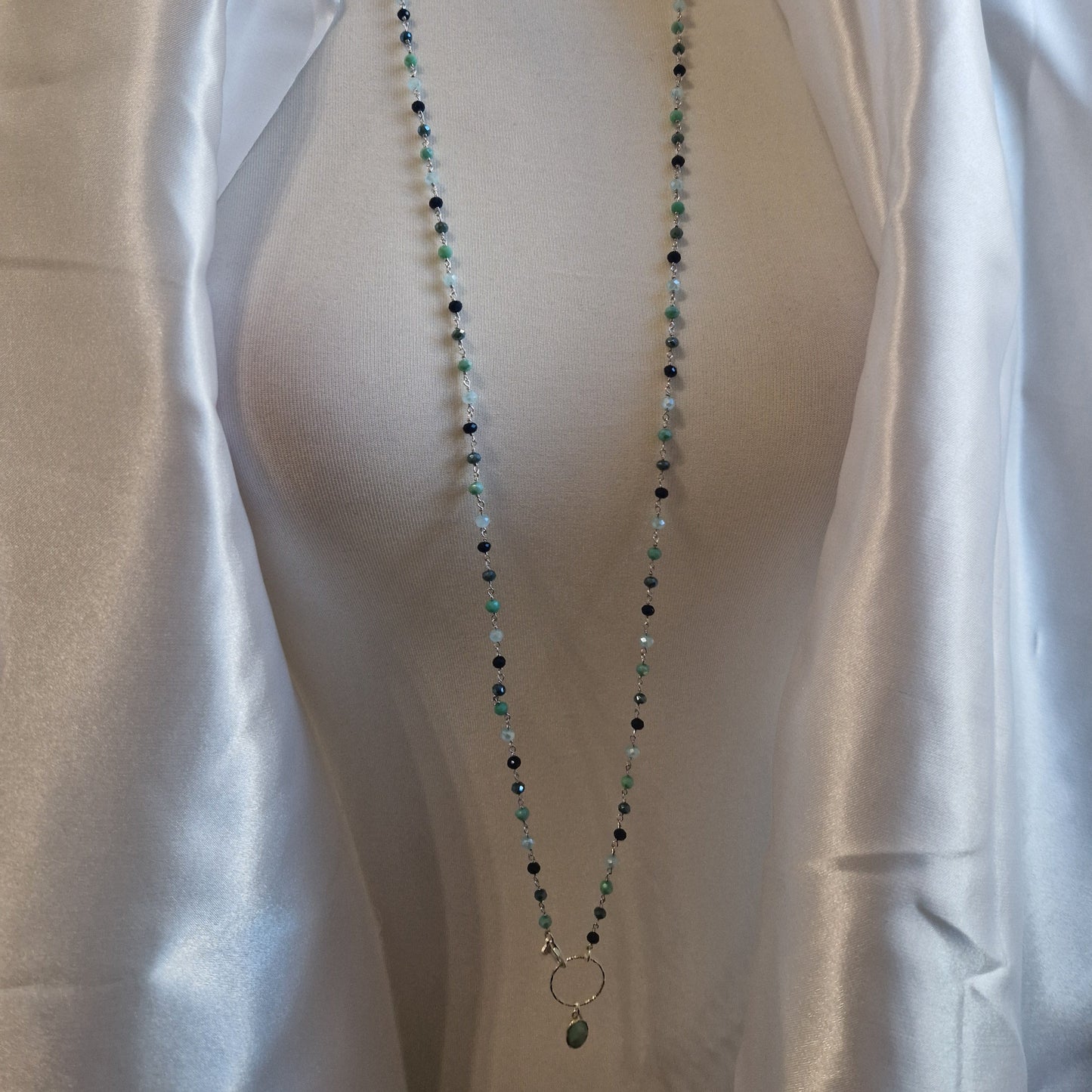 Collana lunga di perline di diverse tonalità di verde e nero, esposta su un manichino con un drappeggio bianco.