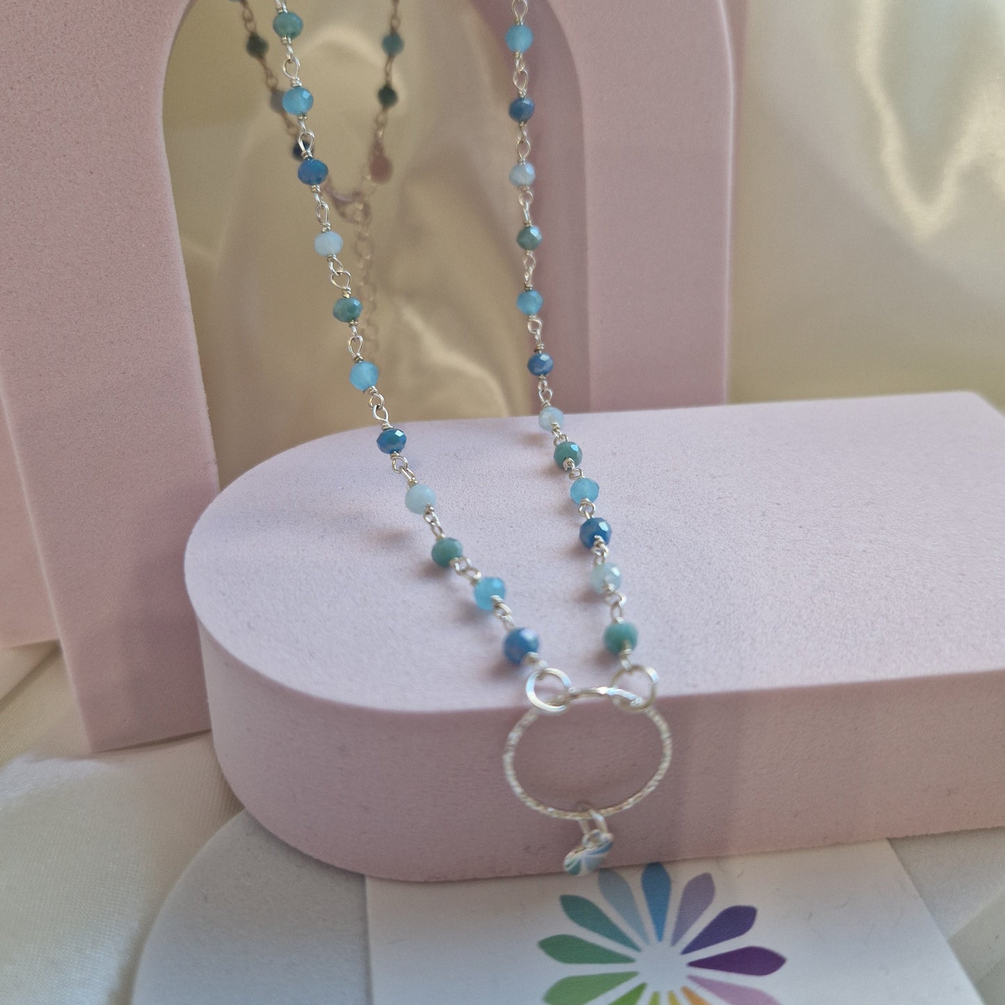 Collana artigianale con piccole perle azzurre e blu, su un supporto rosa.