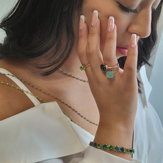 Donna con gioielli in oro rosa, anelli con pietre verdi e nere, bracciale e collana a catena sottile.