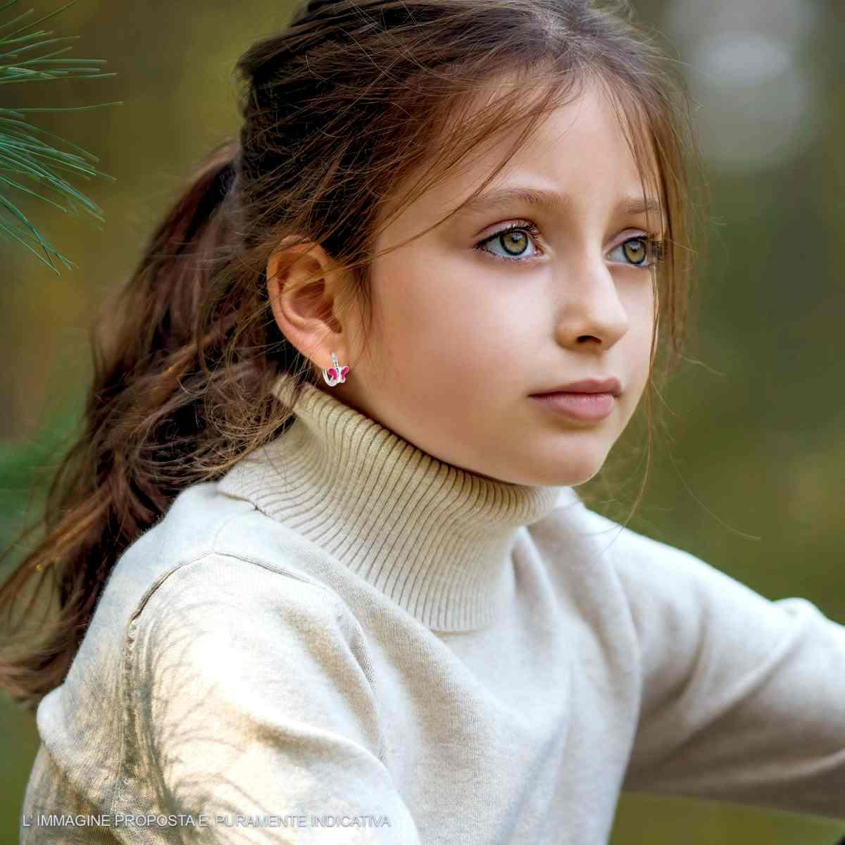 Bambina con un maglione a collo alto e orecchini rosa, seduta all'aperto, guarda lontano.