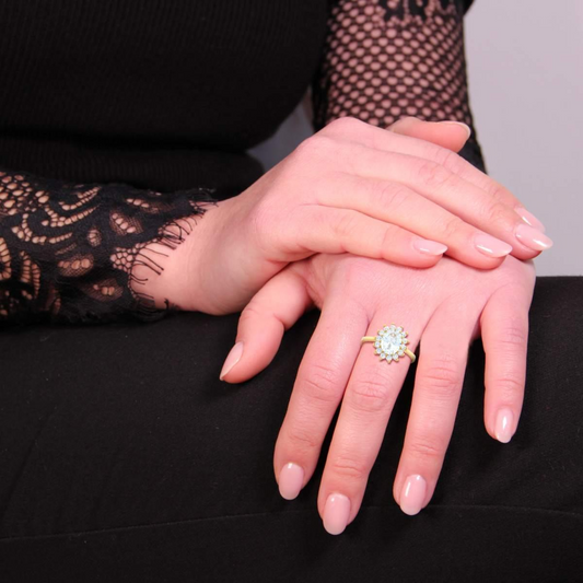 Anello in oro con diamante centrale e contorno di diamanti, indossato su una mano femminile con smalto rosa.