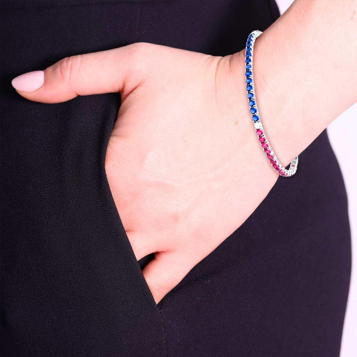 Braccialetto a tennis con pietre blu e rosa, indossato al polso di una donna con pantaloni neri.