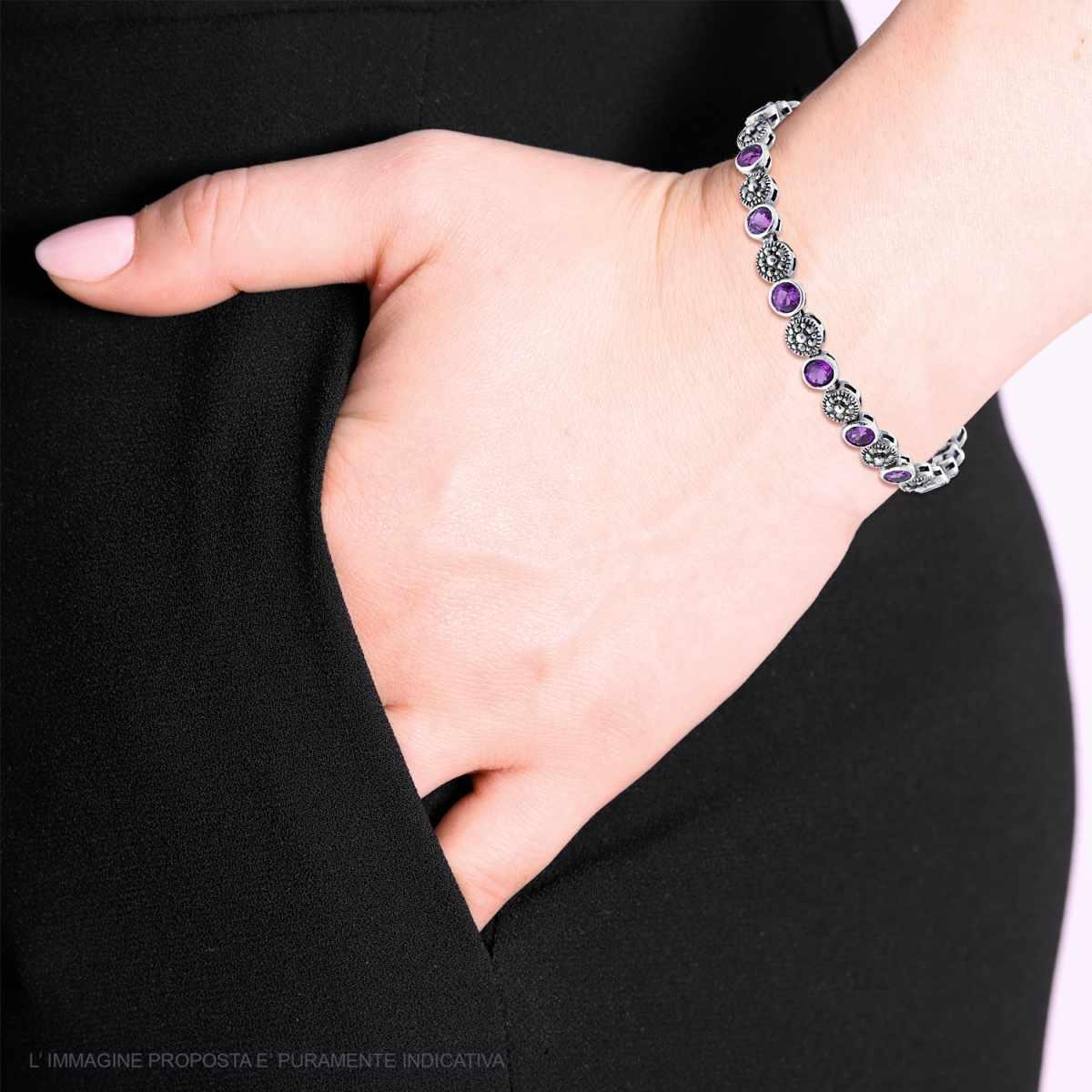 Bracciale tennis in argento con pietre viola indossato al polso di una donna, che ha una mano nella tasca dei pantaloni neri.