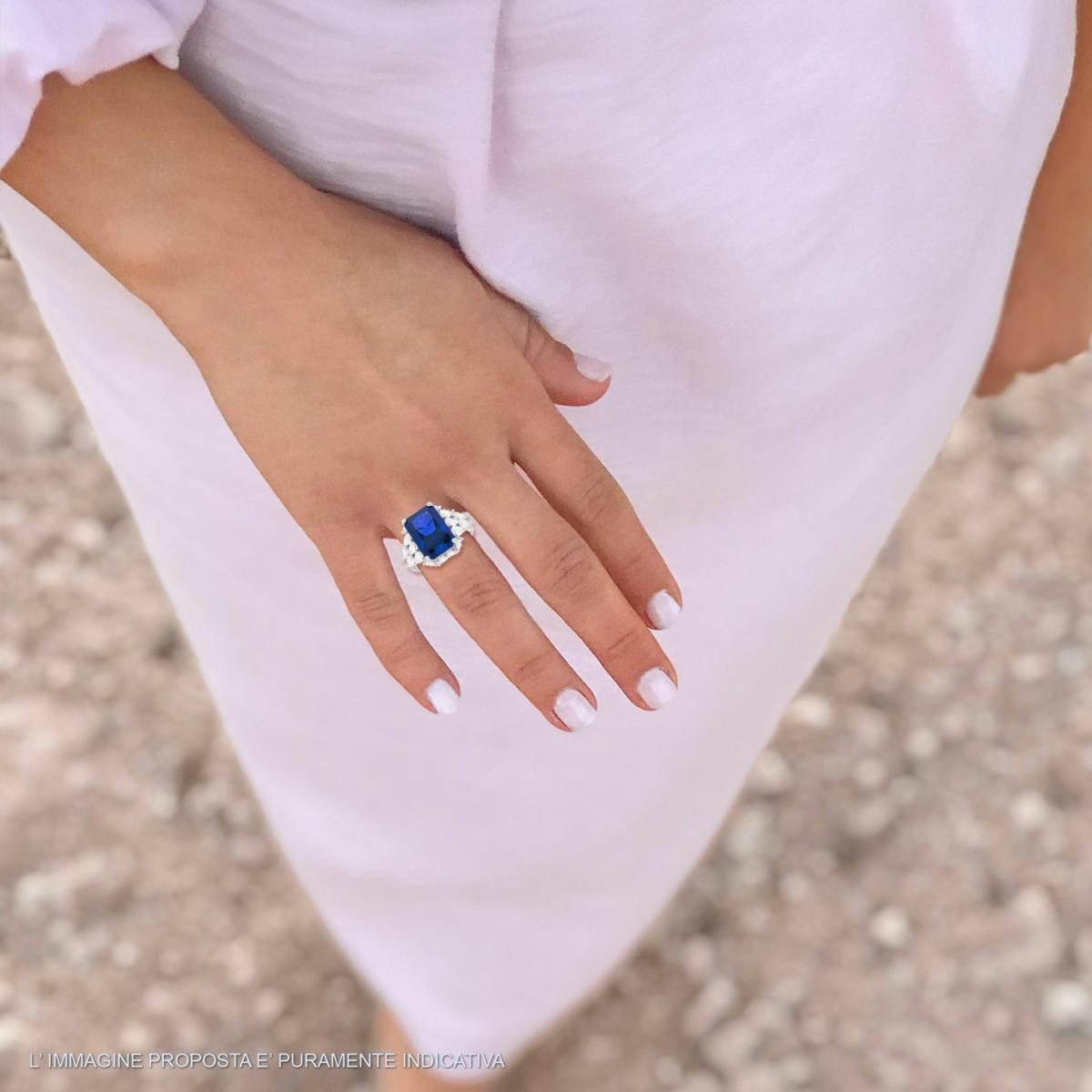 Anello con zaffiro blu e diamanti su una mano con unghie laccate di bianco. L'anello ha una forma rettangolare e una fascia d'argento.