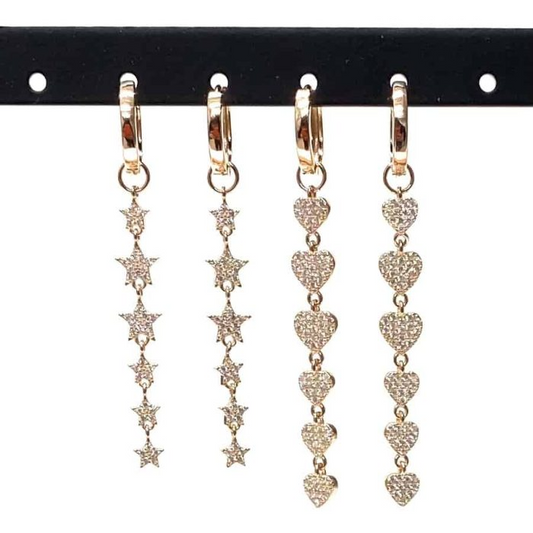 Orecchini pendenti in oro con stelle e cuori, incastonati con piccole gemme scintillanti, esposti su un supporto nero.