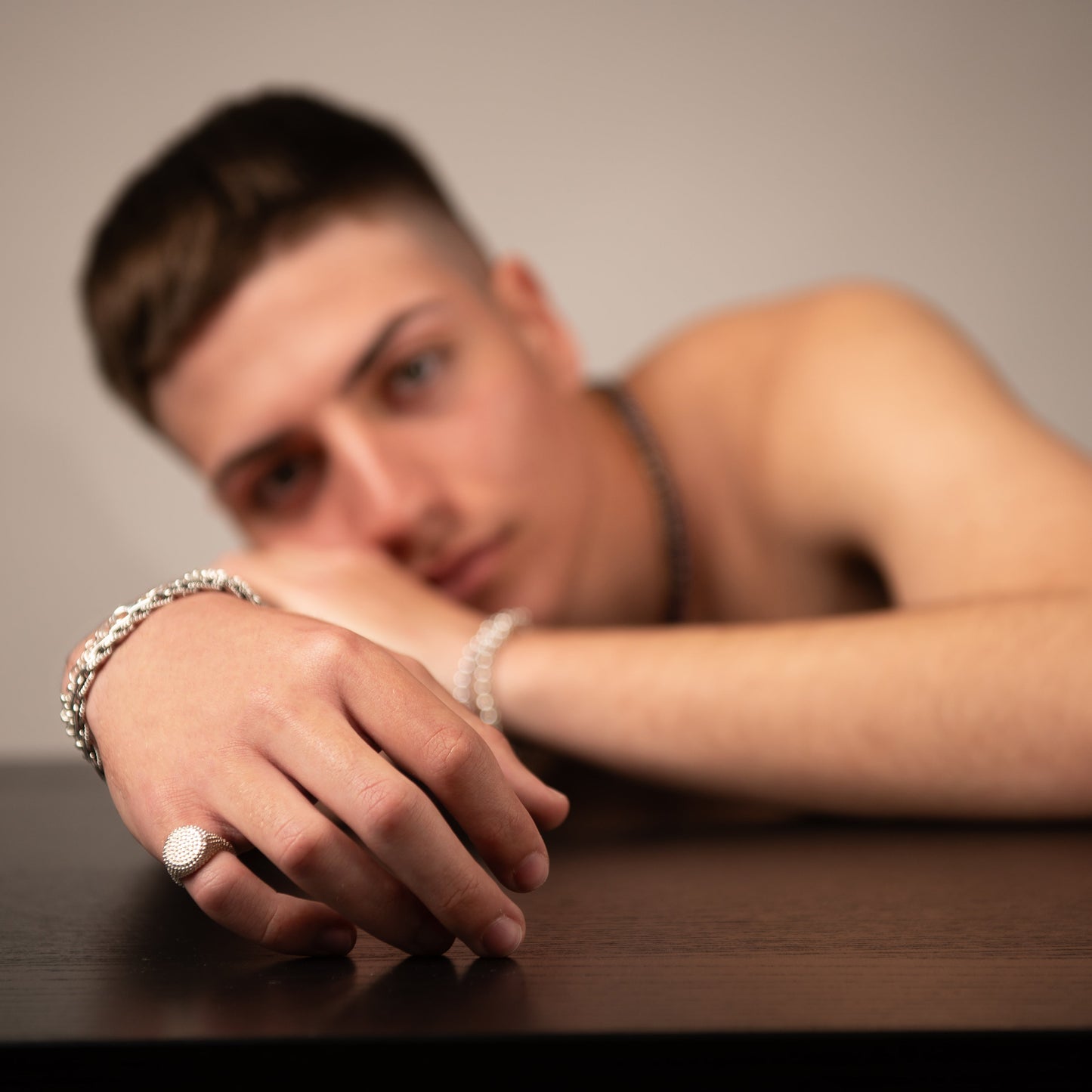 Primo piano di un ragazzo che indossa gioielli in argento, con il braccio e la mano appoggiati su un tavolo scuro.