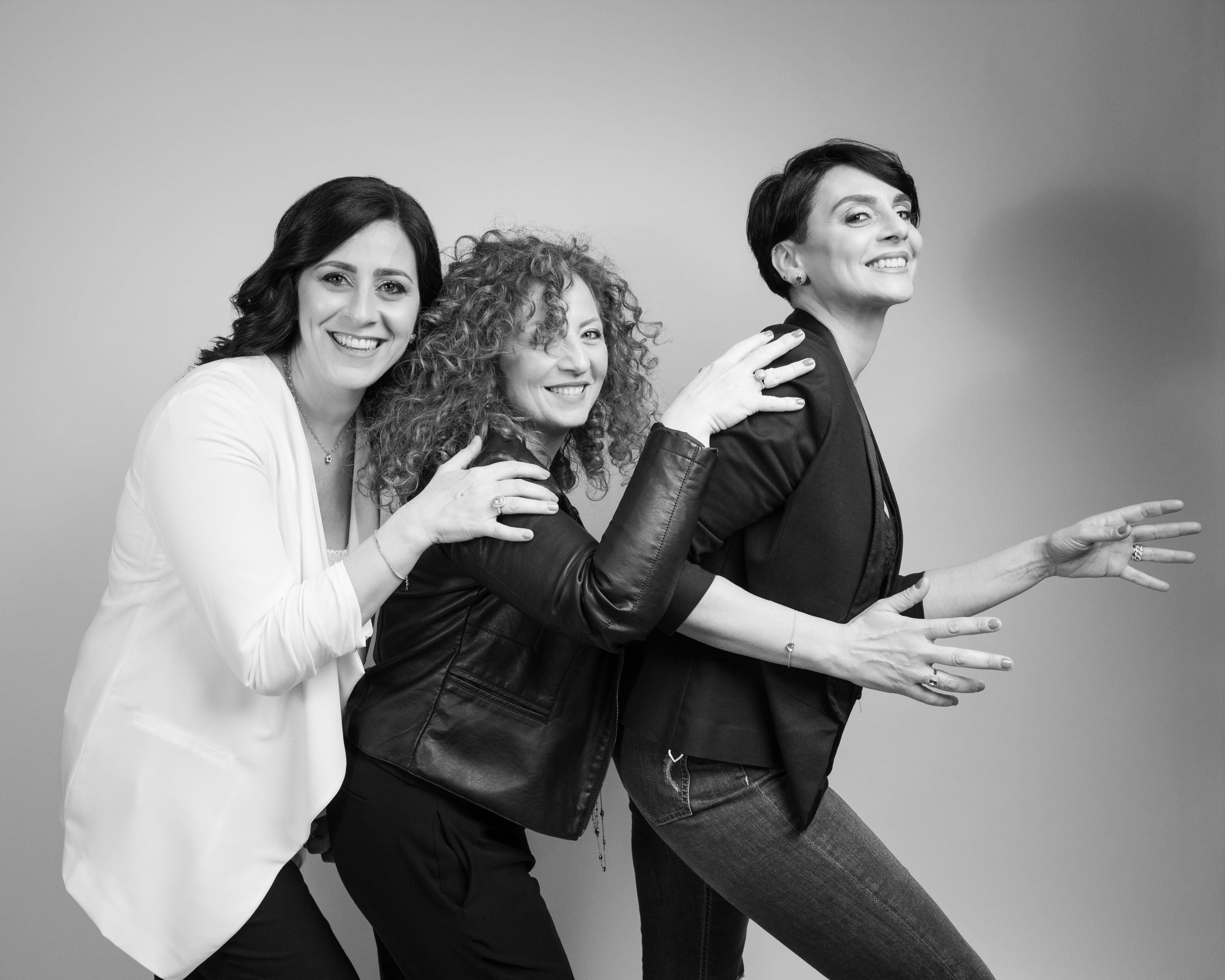Tre donne sorridenti, in fila, si toccano la schiena. La prima, a sinistra, è in primo piano.
