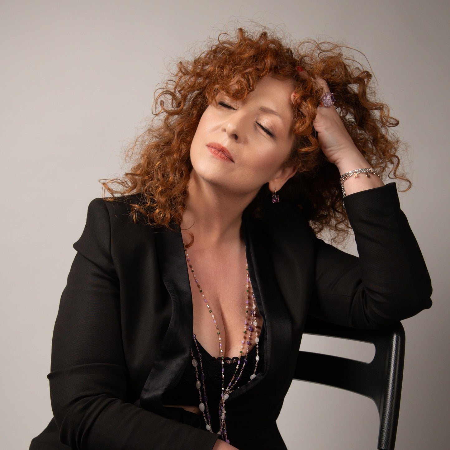 Donna dai capelli ricci e rossi con gli occhi chiusi, che indossa un blazer nero e gioielli, appoggia la mano sulla testa.