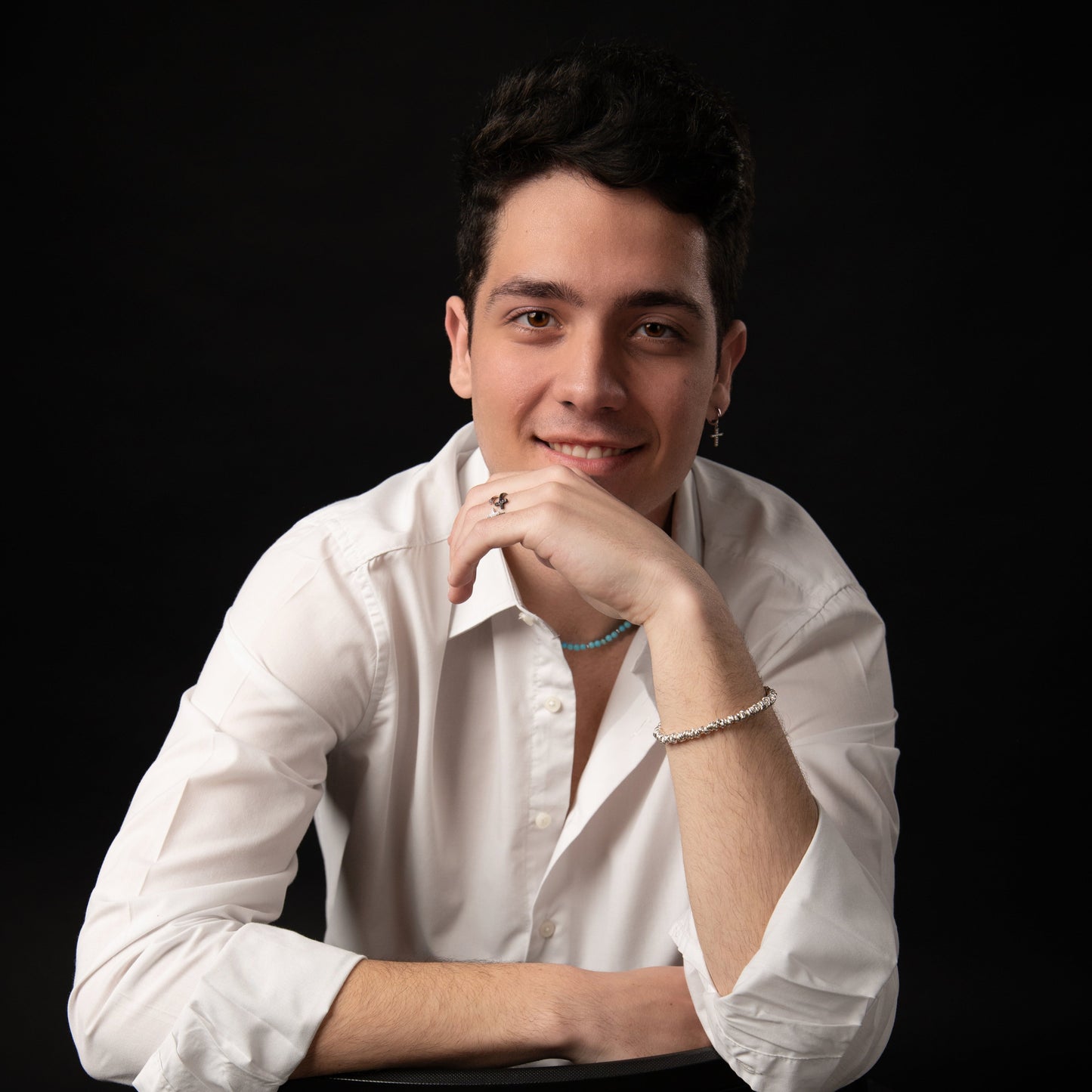 Ritratto in primo piano di un giovane uomo sorridente con camicia bianca e gioielli, che tiene il mento con la mano destra.