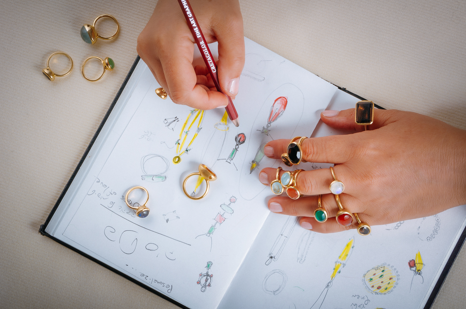 Mani di un designer di gioielli che disegnano schizzi di anelli su un quaderno, con vari anelli d'oro con pietre preziose sparse sulla pagina e indossati. Si evidenzia la maestria artigianale nella creazione di gioielli unici.