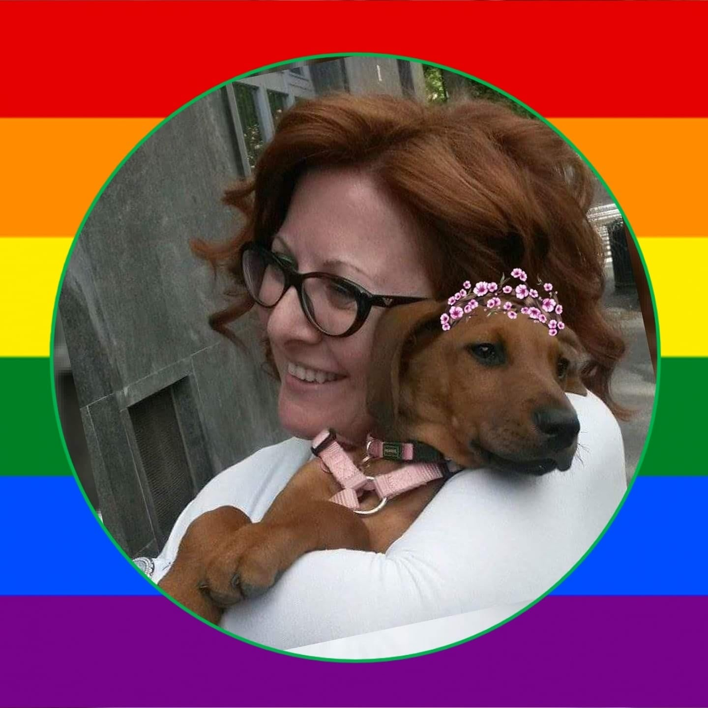 Donna sorridente con occhiali, capelli rossi, abbraccia un cane marrone che indossa una fascia di fiori rosa.