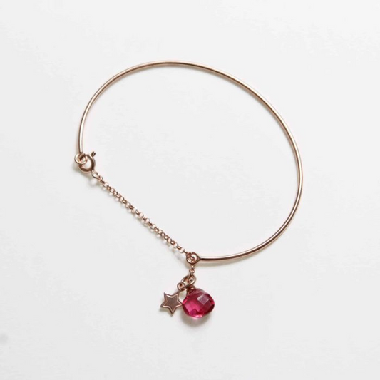 Braccialetto rigido color oro rosa con chiusura a catena e due ciondoli: una stella e una gemma rosa sfaccettata.