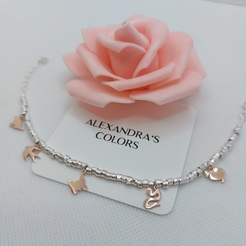 Cavigliera con palline in argento e ciondoli dorati, su sfondo con rosa di schiuma rosa e un cartellino "ALEXANDRA'S COLORS".