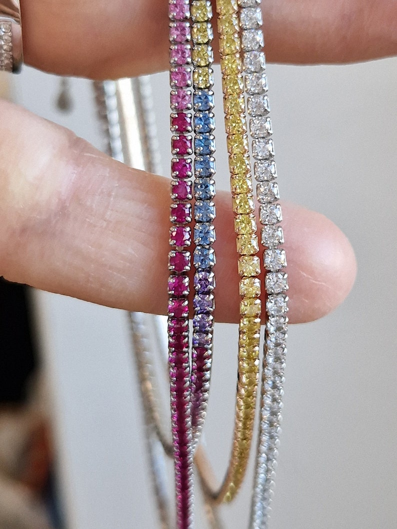 Tre bracciali tennis con gemme colorate: rosa, rubino, zaffiro, gialle e bianche, tenuti tra due dita.