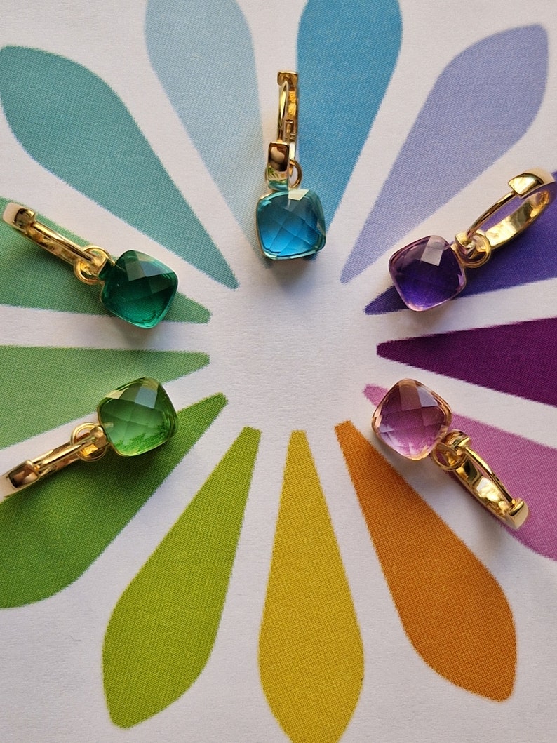 ciondoli dorati con pietre colorate (blu, viola, rosa, giallo e verde) su sfondo con petali arcobaleno.