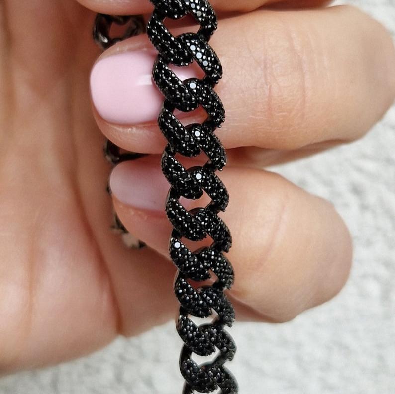 Braccialetto a catena nera, con zirconi incastonati, tenuto in una mano con unghie rosa pallido.