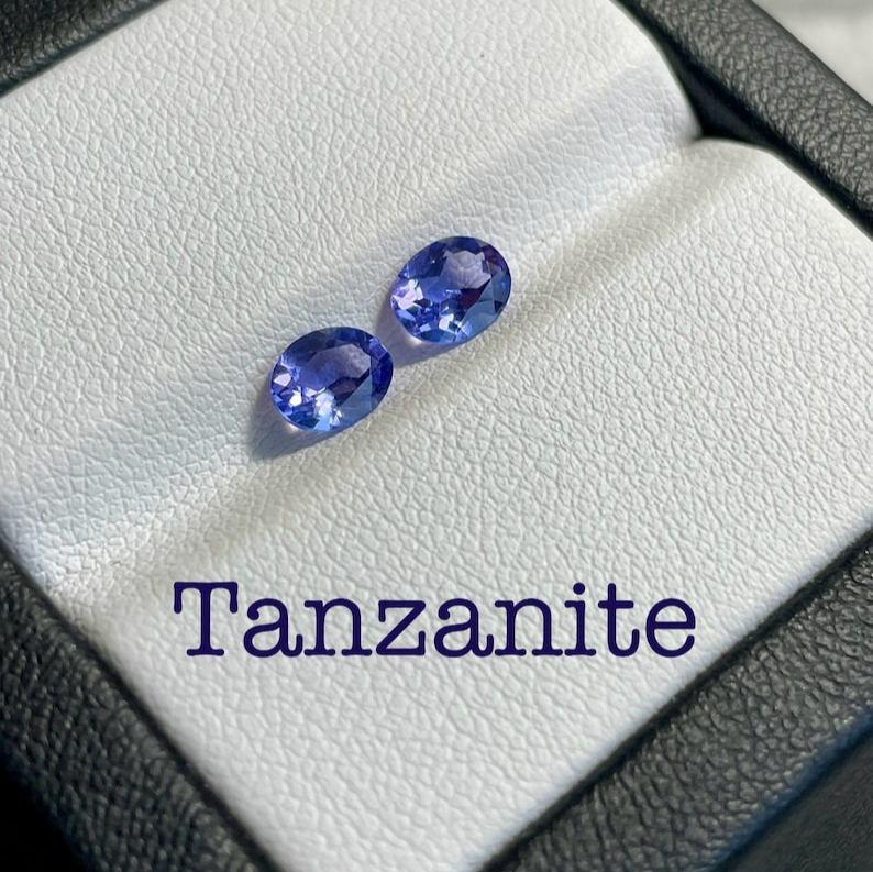 Due gemme ovali di tanzanite viola-blu brillano in un elegante cofanetto bianco con la scritta "Tanzanite" in viola.