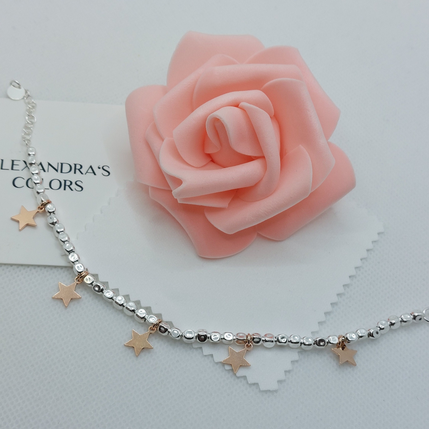 Cavigliera d'argento con perline e ciondoli a stella in oro rosa, su sfondo di carta con scritta "Alexandra's Colors" e una rosa di sapone.