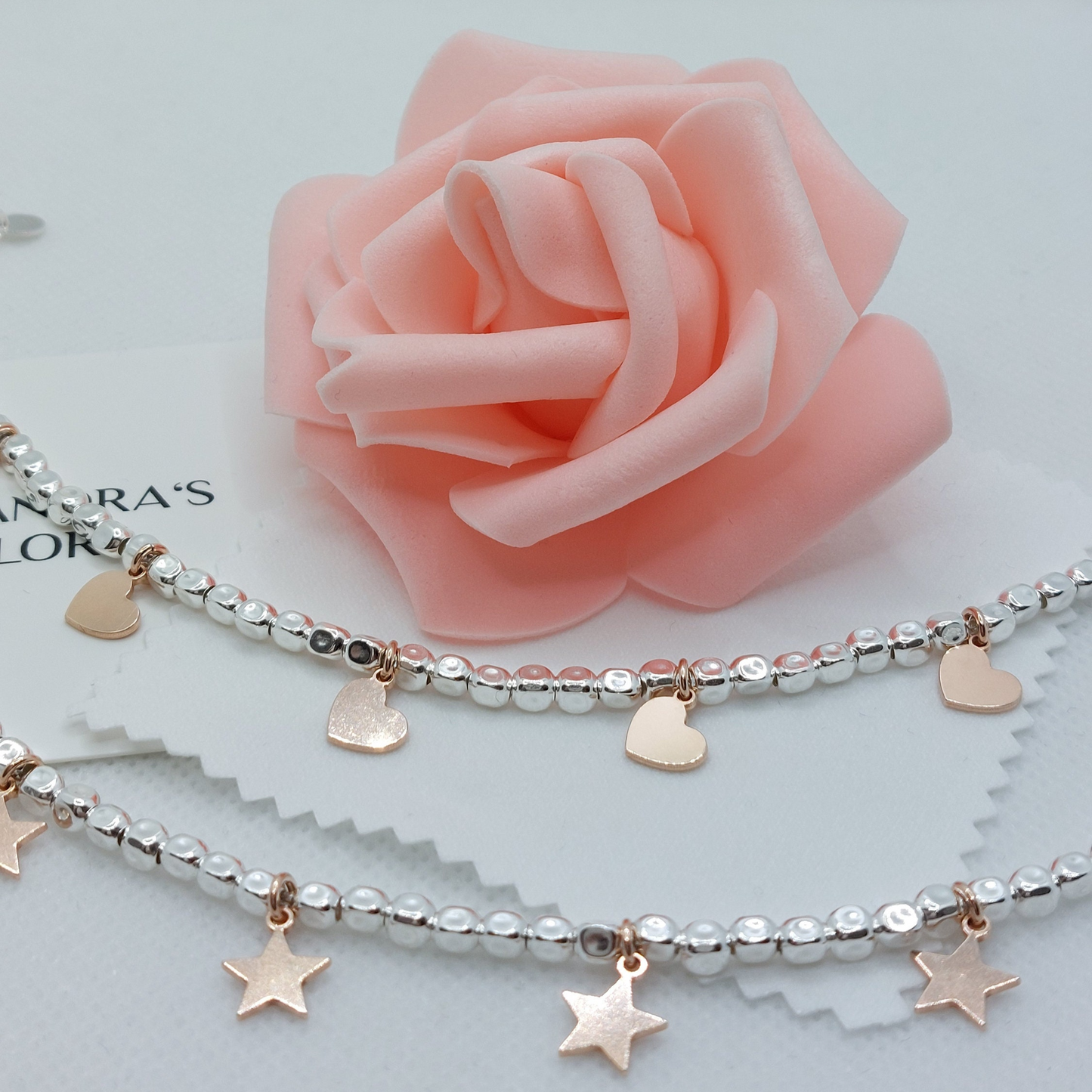 Due bracciali in argento con perline e ciondoli a forma di cuore e stella, su uno sfondo con una rosa rosa.