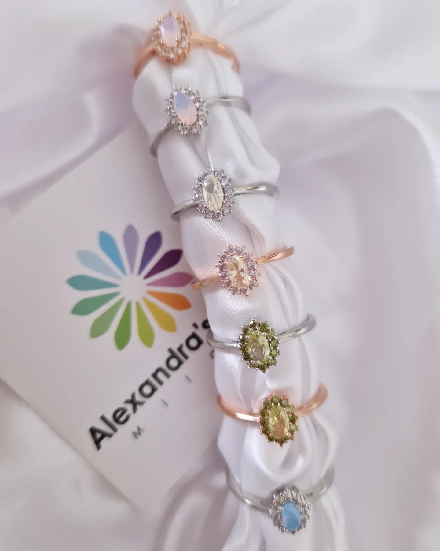 Una linea di sette anelli con gemme di vari colori, su sfondo di tessuto di raso bianco e un logo.
