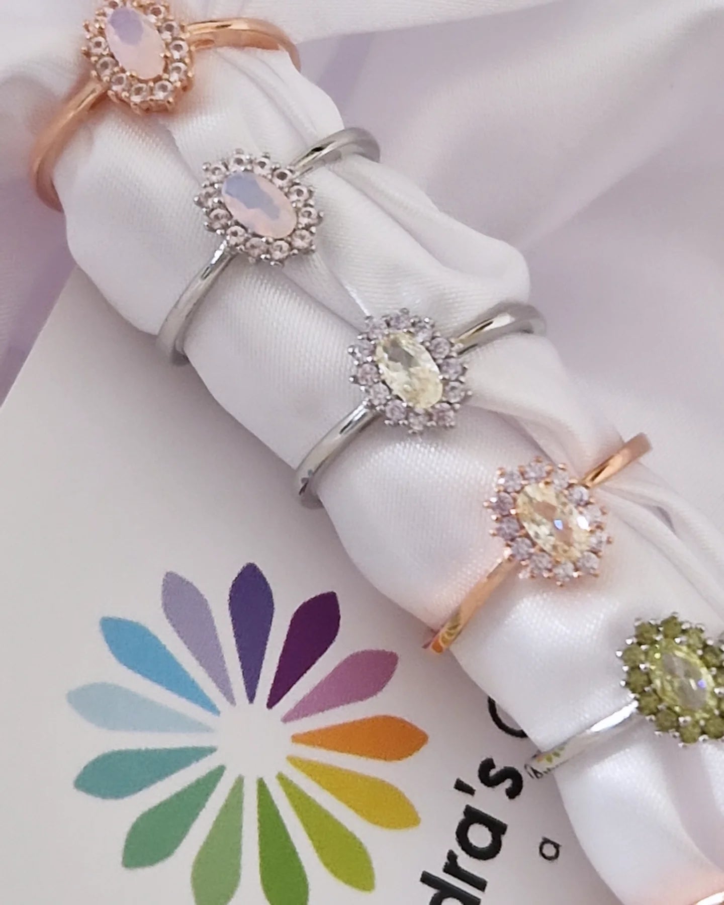 Sei anelli con pietre ovali e brillanti in metallo argento e oro rosa, disposti su un elegante drappeggio di seta bianca.
