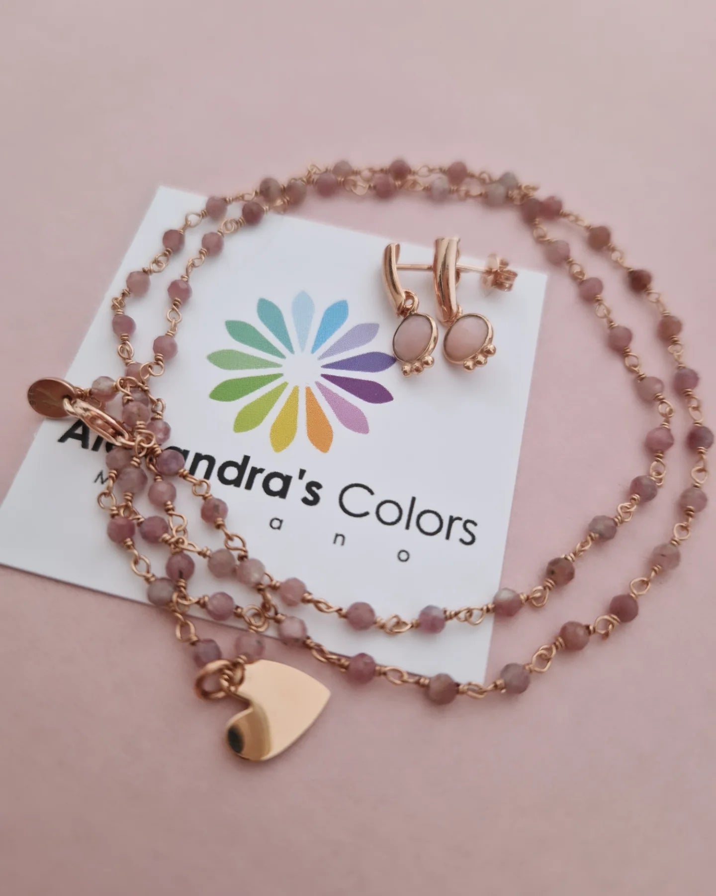 Collana e orecchini in oro rosa con pietre rosa e ciondolo a cuore su biglietto "Alexandra's Colors".