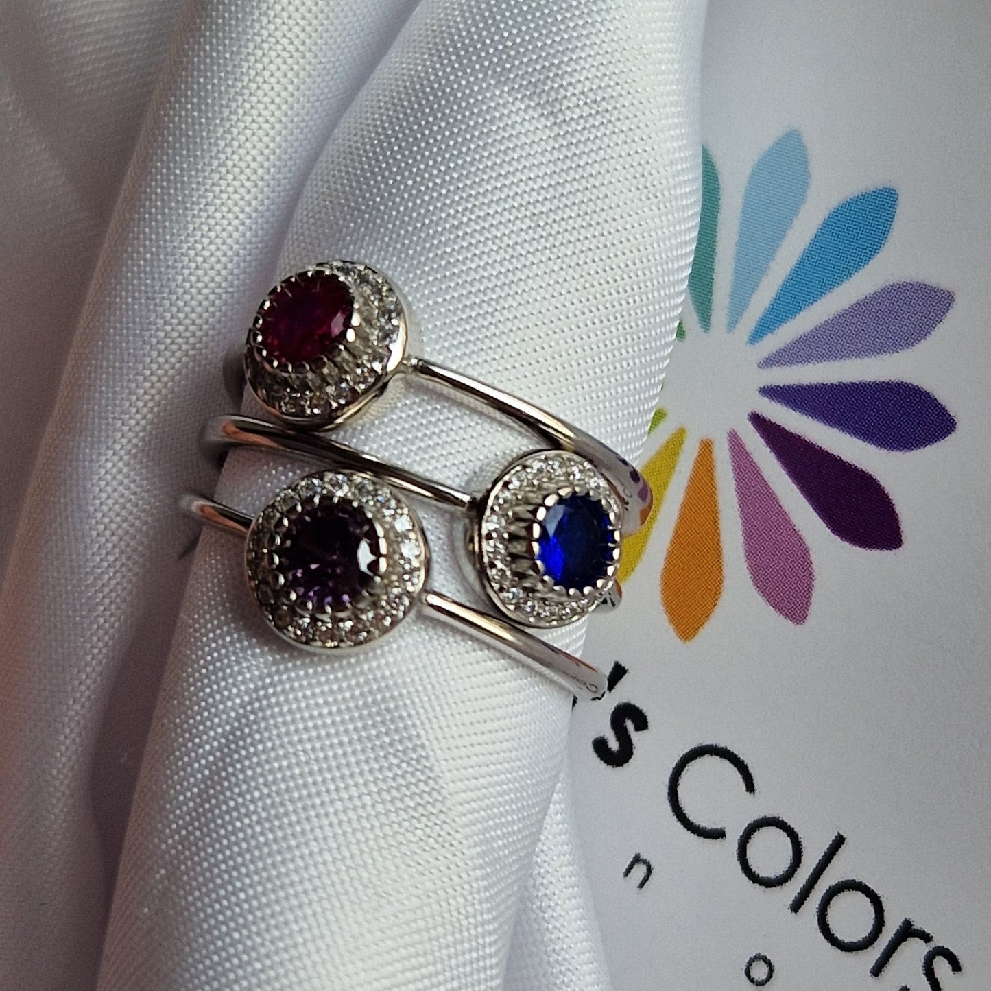 Anelli d'argento con pietre preziose, rosso, viola e blu, avvolti in un tessuto bianco.