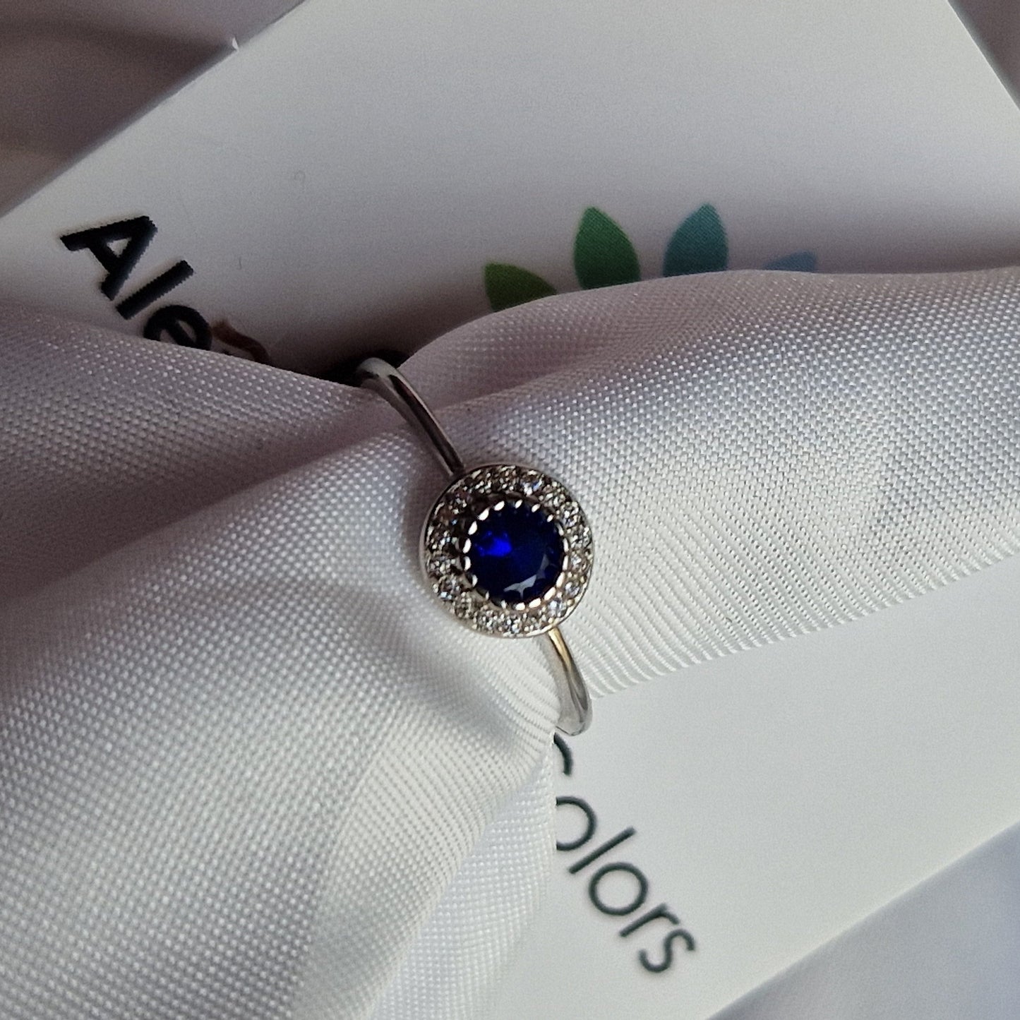 Anello d'argento con zaffiro blu centrale circondato da piccoli diamanti, adagiato su una stoffa bianca.