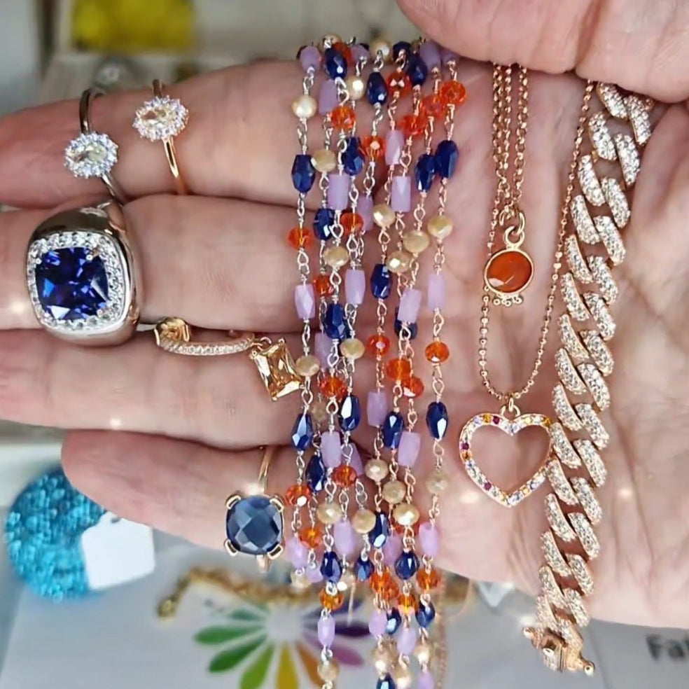Vari gioielli, tra cui anelli con pietre preziose, collane colorate e un bracciale a catena con diamanti.