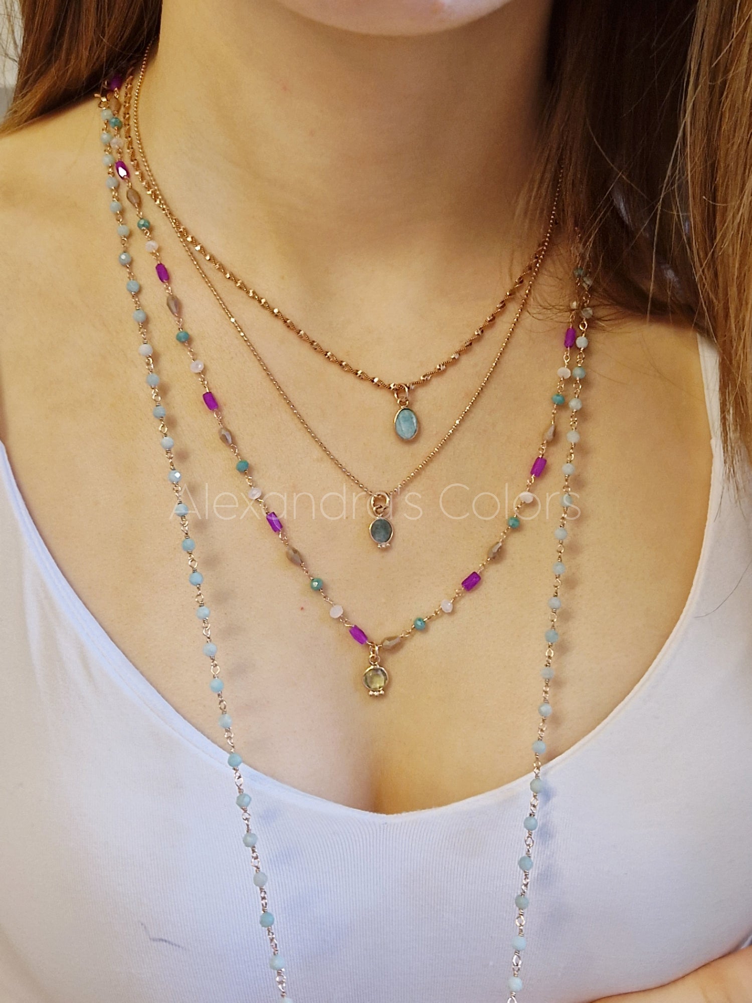 Donna che indossa lunghe collane a strati con gemme colorate nei toni del turchese, del viola e del rosa, con catene d'oro e un ciondolo centrale a forma di goccia, su una canottiera chiara.