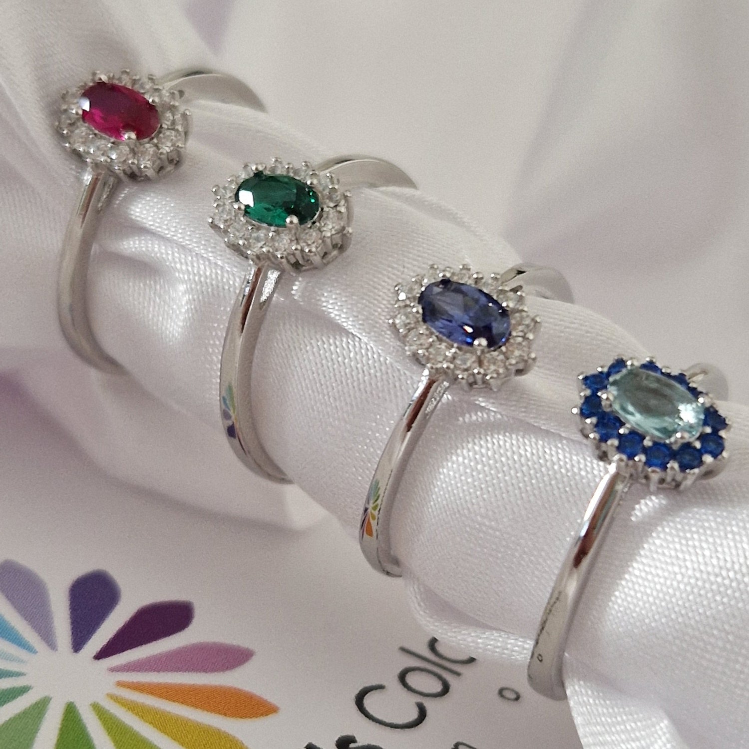 Anelli in argento con pietre preziose: rubino, smeraldo, zaffiro e acquamarina, su un nastro di seta bianca.